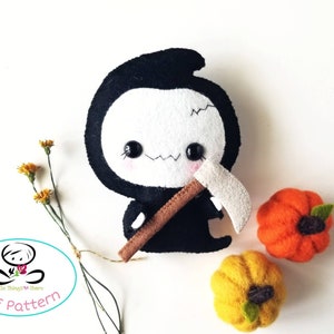 May include: A black felt plush toy of the Grim Reaper with a white face, holding a brown felt scythe. The toy is surrounded by two felt pumpkins, one orange and one yellow.
