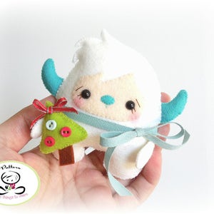 May include: A white felt snowman holding a green felt Christmas tree with red and white buttons. The snowman has blue felt arms and a blue felt bow around its neck.
