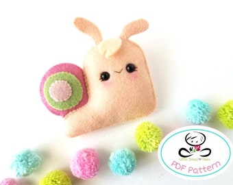 Pode incluir: Um brinquedo de feltro em forma de caracol com uma concha rosa, detalhes verdes e um rosto bonito. O caracol está rodeado de pompons coloridos. A imagem também inclui um logotipo para "Little Things to Share" e o texto "PDF Pattern".