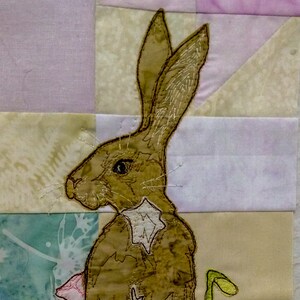 May include: A brown rabbit with white fur on its belly is standing on a patchwork quilt. The rabbit is surrounded by yellow and purple flowers.