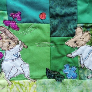 May include: Two embroidered mice playing a game of baseball on a green and brown patchwork quilt. The mice are wearing white shirts and have brown fur. The quilt is decorated with flowers and leaves.