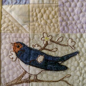 May include: A quilted fabric wall hanging featuring a blue bird embroidered on a branch with white flowers. The background is a patchwork of light and dark fabrics with various quilting patterns.