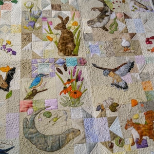 May include: A white quilt with a variety of embroidered animals and plants. The animals include a bat, a squirrel, a bird, a rabbit, a fish, and an otter. The plants include flowers, leaves, and branches.