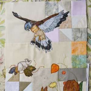 May include: A quilted wall hanging featuring embroidered images of a hawk, a squirrel, a mouse, and various plants. The background is made of different coloured fabric squares.