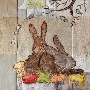 May include: Two brown embroidered bunnies cuddling together on a beige fabric background with green, orange, and red embroidered leaves around them.