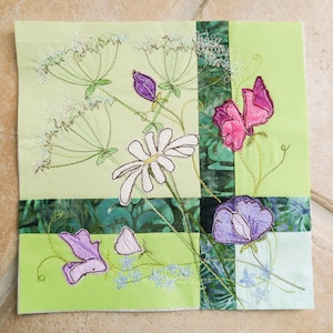 Könnte beinhalten: Ein grüner Stoffquadrat mit gestickten Blumen. Die Blumen sind weiß, lila und rosa. Der Stoff ist in vier Quadrate unterteilt, mit einem grün-weiß gemusterten Stoff in der Mitte.