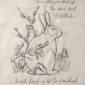 May include: Black and white embroidered illustration of a rabbit with its ears perked up, hiding behind a blackthorn blossom. The text reads "The rabbit pricked up his ears, and I stilled... breath hanging as he crouched behind the blackthorn blossom."