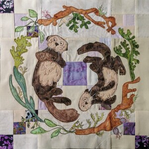 May include: A quilted fabric wall hanging with an embroidered design of two brown sea otters playing in a circle of green and brown foliage. The otters are surrounded by a brown branch with green leaves.