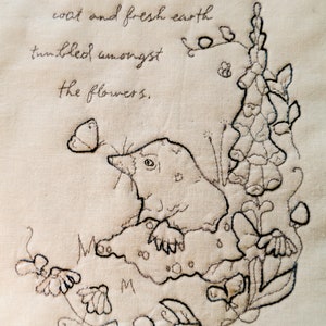 May include: Black and white embroidered illustration of a mole with a butterfly, flowers, and leaves. The text "Sunlight warmed the mole's velvet coat and fresh earth tumbled amongst the flowers." is stitched above the illustration.