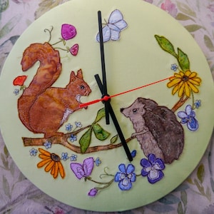 May include: A green clock with a squirrel and a hedgehog embroidered on it. The clock has a black and red hand and is surrounded by flowers and butterflies.