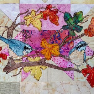 May include: Two bluebirds perched on a branch with brown leaves and red, yellow, and green leaves. The birds are surrounded by a patchwork of pink, white, and brown fabric.