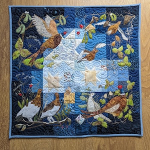 Puede incluir: Una colcha de retazos azul y blanca con un diseño bordado de pájaros y flores. La colcha está hecha de cuadrados y rectángulos, y los pájaros están en varias poses, incluyendo volando, posados y de pie. Las flores son de varios colores, incluyendo rojo, rosa y amarillo.