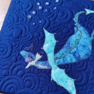 Può includere: Un tessuto trapuntato blu con un motivo di drago ricamato in blu e verde. Il drago sta volando con le ali spiegate e la coda arrotolata. Lo sfondo è blu scuro con un motivo a spirale.