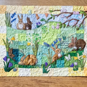 Puede incluir: Un tapiz de pared acolchado con un diseño de patchwork que presenta conejos marrones, hojas verdes y flores amarillas. El fondo es un patrón de cuadros azul claro y blanco.