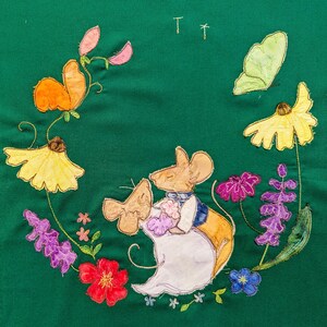 Könnte beinhalten: Ein grüner Stoff mit gestickten Blumen, Schmetterlingen und zwei Mäusen. Die Mäuse halten sich an den Händen und sind in ein weißes Kleid und eine blaue Weste gekleidet. Der Stoff ist mit verschiedenen Blumen verziert, darunter gelbe, lila und rote.