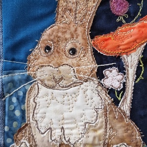 May include: A brown and white fabric bunny rabbit with a white fluffy tail, sitting on a blue background with a mushroom behind it. The bunny has embroidered details and a stitched-on nose.