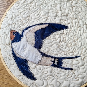 May include: A blue and white embroidered swallow on a cream background with a decorative quilted pattern. The swallow is in flight with its wings spread.