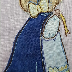 May include: A blue and white fabric mouse wearing a blue cap and a yellow bow. The mouse is holding a yellow ribbon in its right paw.