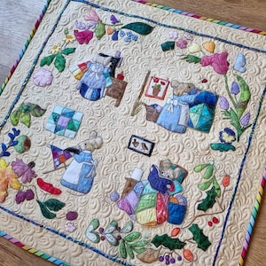 Könnte beinhalten: Ein gesteppter Wandbehang mit einer skurrilen Szene mit mehreren Mäusen, die Kleidung tragen und von Blumen, Blättern und anderen naturinspirierten Elementen umgeben sind. Der Hintergrund ist cremefarben mit einem gesteppten Muster.