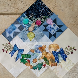 May include: A white fabric square with a blue and white star pattern, a blue and white floral pattern, and a brown squirrel with a blue bow. The fabric square is surrounded by a white border.