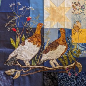 May include: A colorful patchwork quilt with a star design in the center. Two embroidered birds are perched on a branch with green leaves. The birds are white with brown accents and have long beaks. The quilt is made of various fabrics, including blue, white, yellow, and brown.