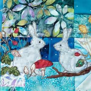 May include: Two white rabbits embroidered on a blue and green patchwork quilt. The rabbits are facing each other and are surrounded by flowers and leaves.