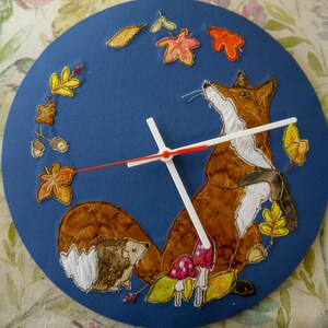 May include: A round, blue clock featuring an embroidered fox, hedgehog, and autumn leaves. The fox is brown and white, looking upwards. The clock hands are white with a red second hand. The design includes colorful leaves and mushrooms.