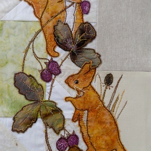 May include: Two orange squirrels embroidered on a white fabric background. The squirrels are climbing a vine with green leaves and purple berries. The background is a patchwork of different fabrics.