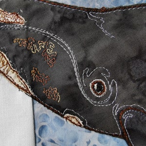 May include: Close-up of a dark gray fabric whale with brown and white embroidery. The whale's eye is outlined in white and filled with a black button.