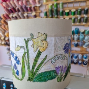May include: A beige lampshade with an embroidered floral design featuring yellow daffodils, bluebells, and purple crocuses. The design is on a white background with a subtle pattern.