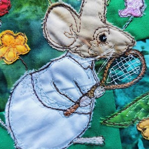 Könnte beinhalten: Eine weiße Maus, die ein blaues Hemd trägt und einen braunen Tennisschläger hält. Die Maus ist auf einem grünen Stoffhintergrund mit gelben und rosa Blumen gestickt.