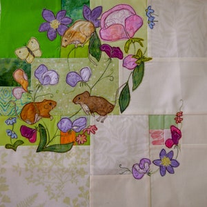 Könnte beinhalten: Eine Patchwork-Decke mit floralem und tierischem Thema. Die Decke zeigt gestickte Bilder von Mäusen, Schmetterlingen und Blumen in Rosa-, Grün- und Lilatönen.