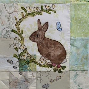 May include: A brown rabbit sits on a green leafy branch with white flowers. A blue butterfly is flying above the rabbit. The background is a patchwork of green and white fabric.