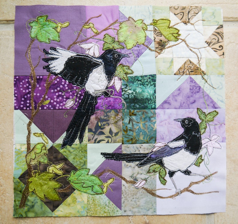 May include: A quilted fabric wall art featuring two black and white magpies perched on a branch with green leaves. The background is a patchwork of purple, green, and white fabric squares.