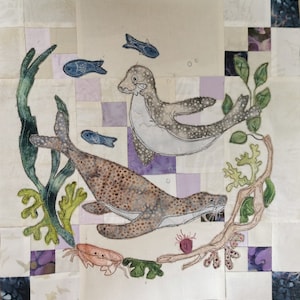 May include: A quilted fabric wall art featuring a brown and white seal swimming with a smaller gray seal. The seals are surrounded by green seaweed and blue fish. The background is a patchwork of purple, white, and gray squares.