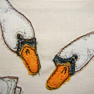 Könnte beinhalten: Nahaufnahme von zwei gestickten weißen Schwänen mit orangefarbenen Schnäbeln und schwarzen Augen. Die Schwäne sind auf einem weißen Leinwandgrund gestickt.