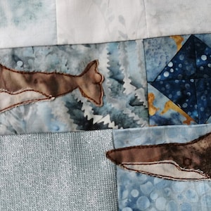 May include: Two brown and white whale appliques on a blue and white fabric background. The whales are stitched with a white thread and have a light brown body with a white belly.