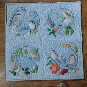 May include: A light blue quilted wall hanging with embroidered birds on branches, flowers, and leaves. The birds are in various colours, and the quilt has a decorative stitched pattern. The overall theme is nature and birds.