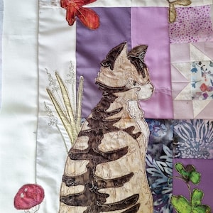 May include: A fabric art piece depicting a cat with brown stripes, set against a patchwork background of purple, white, and floral patterns. Appliqued leaves, mushrooms, and foliage add detail to the design.