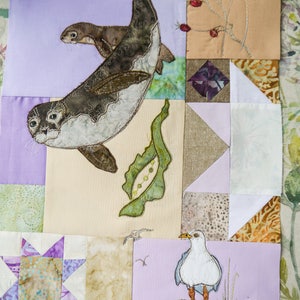 Könnte beinhalten: Eine bunte Patchwork-Decke mit verschiedenen gestickten Motiven, darunter eine Möwe, zwei Robben und ein Blumenmotiv. Die Decke besteht aus verschiedenen Stoffen in den Farben Lila, Braun, Beige und Weiß.
