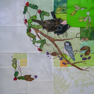 Könnte beinhalten: Eine weiße Stoffdecke mit Patchwork-Muster, auf der gestickte Vögel und Stechpalmenblätter zu sehen sind. Die Vögel sind schwarz und blau, die Stechpalmenblätter grün mit roten Beeren.