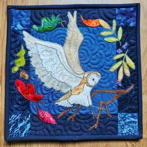 Könnte beinhalten: Ein quadratisches Wandbehang aus Quiltstoff mit einer Schleiereule mit ausgebreiteten Flügeln, die auf einem Ast sitzt. Die Eule ist cremefarben und braun, vor einem blauen Hintergrund mit Blatt- und Eicheldekorationen. Dunkelblauer Rahmen.