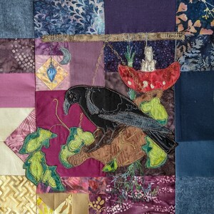 以下が含まれることがあります： パッチワークのキルトに、赤いキノコと緑の葉が刺繍された黒いカラス。キルトは青、紫、ピンク、茶色の様々な生地で作られています。