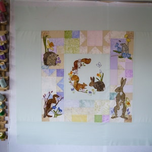 May include: A handmade quilt with embroidered woodland animals, including squirrels, rabbits, and a hedgehog, set within a patchwork design. The quilt is displayed against a light green backdrop. The quilt's dimensions are in centimeters.