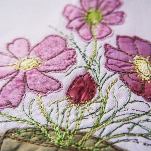 May include: Close-up of a hand-stitched embroidery design featuring pink and purple flowers with green stems on a white fabric background.