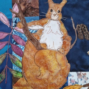 May include: A fabric art piece featuring a squirrel perched on a large, textured, orange-brown shell. The squirrel has brown fur and a white chest. The background is a patchwork of blue, brown, and patterned fabrics, with stitched details.