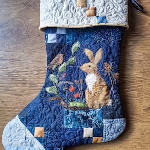 May include: A blue and white quilted Christmas stocking with a brown rabbit and a red bird applique. The stocking has a white cuff with a yellow and brown patchwork design.