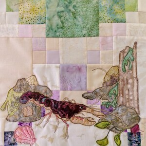 May include: A close-up of a quilted fabric artwork depicting a crayfish on a rock with seaweed and a log. The artwork is made with various shades of brown, green, and purple fabric.