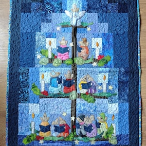 May include: A rectangular fabric quilt featuring a dark blue background with a central tree design. The tree is decorated with appliqued mice wearing colourful clothing, holding books, and standing near white candles with yellow flames and stars.