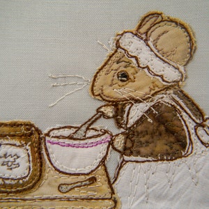 May include: A fabric applique featuring a mouse wearing a bonnet and apron, stirring a bowl. The mouse is brown and tan, with white whiskers and a white apron. A jar and spoon are also visible. The background is a light blue.